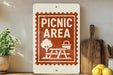 a sign that reads "Picnic Area" with a picnic table and basket icon, placed on a shelf next to a wooden cutting board and some potted plants.