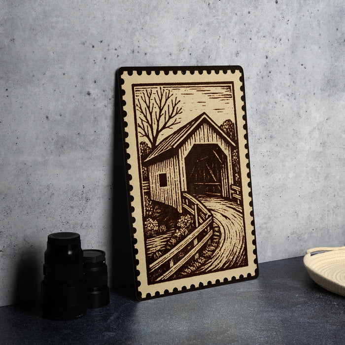 A framed illustration of a covered bridge is displayed on a table, accompanied by a stack of black camera lenses and a white dish.