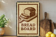 A breadboard sign with a loaf of bread on it, placed on a wooden surface next to kitchen utensils and a potted plant.