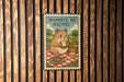 A metal sign with a cartoon hamster sitting on a picnic blanket, holding a sandwich and a drink, with the text "HAMSTE AT PICNIC" displayed on it.