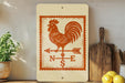 a wooden board with a rooster and a compass rose design, placed on a shelf next to a wooden cutting board and some lemons.