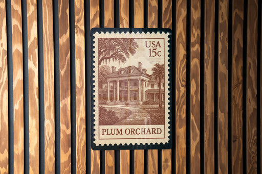 A US postage stamp featuring an illustration of a building, likely a house or mansion, with the text "PLUM ORCHARD" printed on it. The stamp is affixed to a wooden wall with vertical slats.