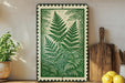 a framed illustration of green ferns on a shelf, with a wooden cutting board and a potted plant to the left.