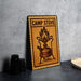 A metal sign with a camp stove illustration on it, placed on a countertop next to some black camera lenses.