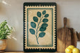a framed print of a plant with green leaves, placed on a shelf next to a wooden cutting board and a potted plant.