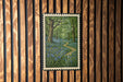 A framed painting depicting a lush forest scene with a winding path through the trees and blue flowers.