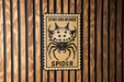 a sign with a spider on it, mounted on a wooden wall.