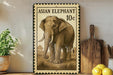 a framed illustration of an elephant, which appears to be a postage stamp-like design. The elephant is depicted in a detailed, realistic style, and the stamp-like border suggests it is a reproduction of a postage stamp. The elephant is standing on a grassy area, and there are some potted plants and kitchen utensils visible in the background.