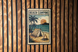 A vintage-style metal sign with a beach camping scene, featuring a tent, palm trees, and a campfire, is displayed on a wooden wall.