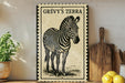 a framed illustration of a zebra, with a wooden cutting board and a potted plant in the background.