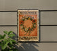 A vintage-style metal sign with a floral wreath design hangs on a gray wall, accompanied by a small green plant to the left.
