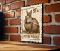 A wooden table with a brick wall in the background, and a rectangular metal sign featuring a rabbit illustration and the text "30c JERSEY WOOLY" hanging on the wall.