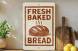a sign that reads "Fresh Baked Bread" with a picture of a loaf of bread on it. The sign is placed on a shelf next to a wooden cutting board and a potted plant.