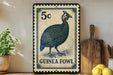 A decorative metal sign featuring a detailed illustration of a guinea fowl, with the text "GUINEA FOWL" printed on it. The sign is placed on a shelf next to a wooden cutting board and a potted plant.
