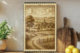 a framed illustration of a rural landscape with a winding road, a church, and a village in the background.
