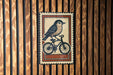 A vintage-style metal stamp depicting a bird riding a bicycle is displayed on a wooden wall.