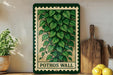 A framed print of a lush green plant with large leaves is displayed on a shelf, accompanied by a wooden cutting board, a potted plant, and some lemons.