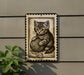 A framed illustration of a cat holding a ball of yarn is displayed on a wall, with a plant visible to the left.