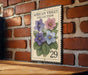 A decorative African violet postage stamp is displayed on a brick wall, featuring a vibrant illustration of purple, blue, and pink flowers.