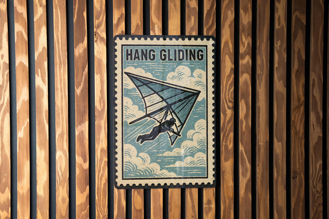 A vintage-style metal sign with the text "HANG GLIDING" and an illustration of a person hang gliding against a cloudy sky background.