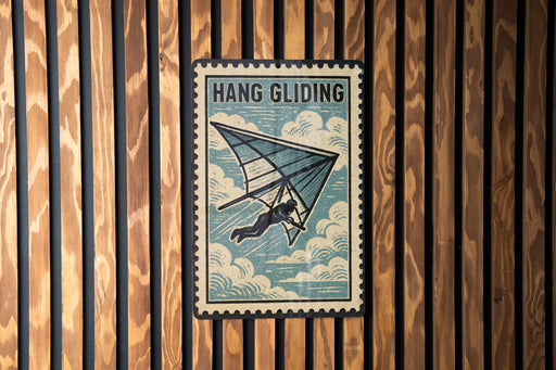 A vintage-style metal sign with the text "HANG GLIDING" and an illustration of a person hang gliding against a cloudy sky background.
