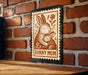 A wooden table with a brick wall in the background, and a framed stamp-like artwork depicting a mother rabbit and her baby rabbit.