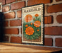 A metal sign with a stamp-like border hangs on a brick wall, featuring a vibrant illustration of orange marigold flowers.