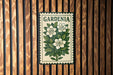 A vintage-style metal sign with the word "GARDENIA" displayed prominently in the center, featuring a floral illustration of white flowers and green leaves.