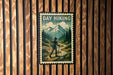 A vintage-style metal sign with the text "DAY HIKING" and an illustration of a hiker walking on a trail through a mountainous landscape.