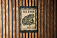 A sign with a stamp-like design featuring a frog, mounted on a wooden wall.