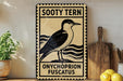 A vintage-style metal sign with a black and white illustration of a bird, labeled "Sooty Tern" and "Onychophron Fuscatus", is displayed on a wooden shelf next to a potted plant and a cutting board.