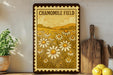 A framed print of a field of flowers is displayed on a shelf, with a wooden cutting board and a potted plant to the left.