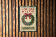 A vintage-style holiday wreath with a red ribbon and greenery is displayed on a wooden wall.