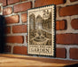 A metal sign with a stamp-like design hangs on a brick wall, depicting a formal English garden.