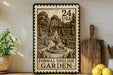 a large stamp-like artwork depicting a formal English garden, with a fountain and trees in the center. The stamp has a value of 24 US dollars and is displayed on a wooden shelf next to a potted plant, a cutting board, and some lemons.