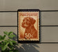 A postage stamp featuring a dog's head is mounted on a wall, with a plant visible to the left.