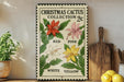 A Christmas cactus collection sign with illustrations of red, pink, white, and yellow flowers, displayed on a wooden shelf next to a potted plant and a wooden cutting board.