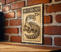 A metal sign with a gecko illustration and the price of 25 cents displayed on a brick wall.