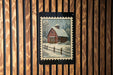 A framed painting of a red barn with a snow-covered roof is displayed on a wooden wall.