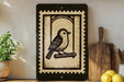 a black and white stamp-like artwork depicting a bird perched on a branch, placed on a shelf next to a wooden cutting board and a potted plant.