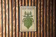 A metal sign with a leaf insect illustration is mounted on a wooden wall.