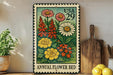 A colorful metal stamp depicting a variety of flowers, including daisies, chrysanthemums, and other blooms, is displayed on a shelf next to a wooden cutting board and a potted plant.