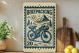 a wooden sign with a bicycle and mountain scenery, along with a stamp-like design, placed on a wooden surface next to a potted plant and some kitchen utensils.