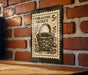 A vintage-style foraging basket postage stamp is displayed on a brick wall, with a wooden table below it.