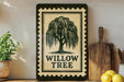a decorative sign with a willow tree design, placed on a shelf next to a wooden cutting board and some kitchen utensils.