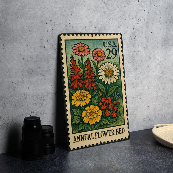 A metal stamp depicting a colorful floral arrangement, with the text "Annual Flower Bed" printed on it, is placed on a table next to some black camera lenses.