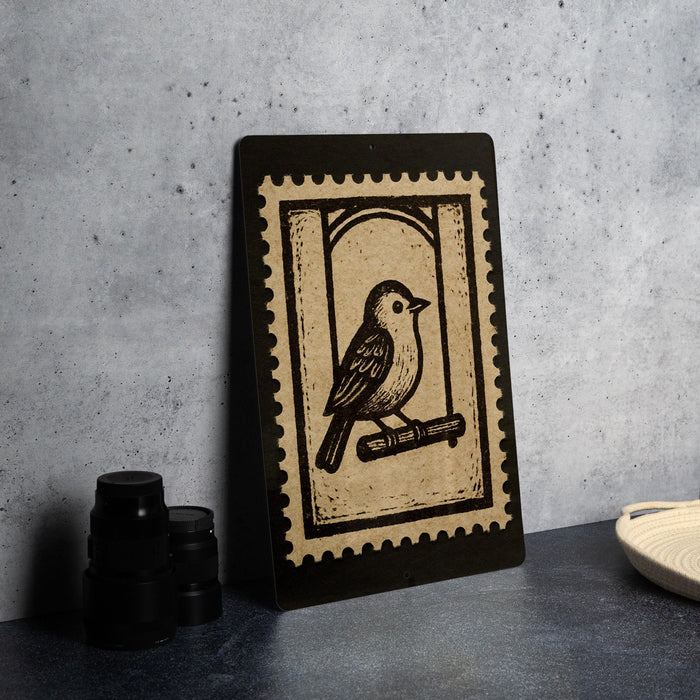 a black and white stamp-like artwork depicting a bird perched on a branch, placed on a dark surface next to some black camera lenses.