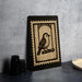 a black and white stamp-like artwork depicting a bird perched on a branch, placed on a dark surface next to some black camera lenses.