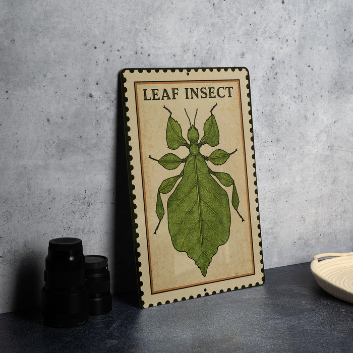 A metal sign with a leaf insect illustration on it, placed on a table next to some black camera lenses.