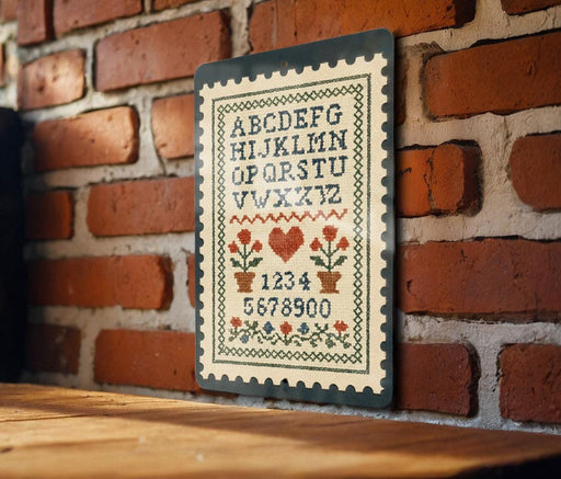 a cross-stitch pattern on a wooden surface, with a brick wall in the background. The pattern includes the letters of the alphabet and numbers, as well as various floral and geometric designs.